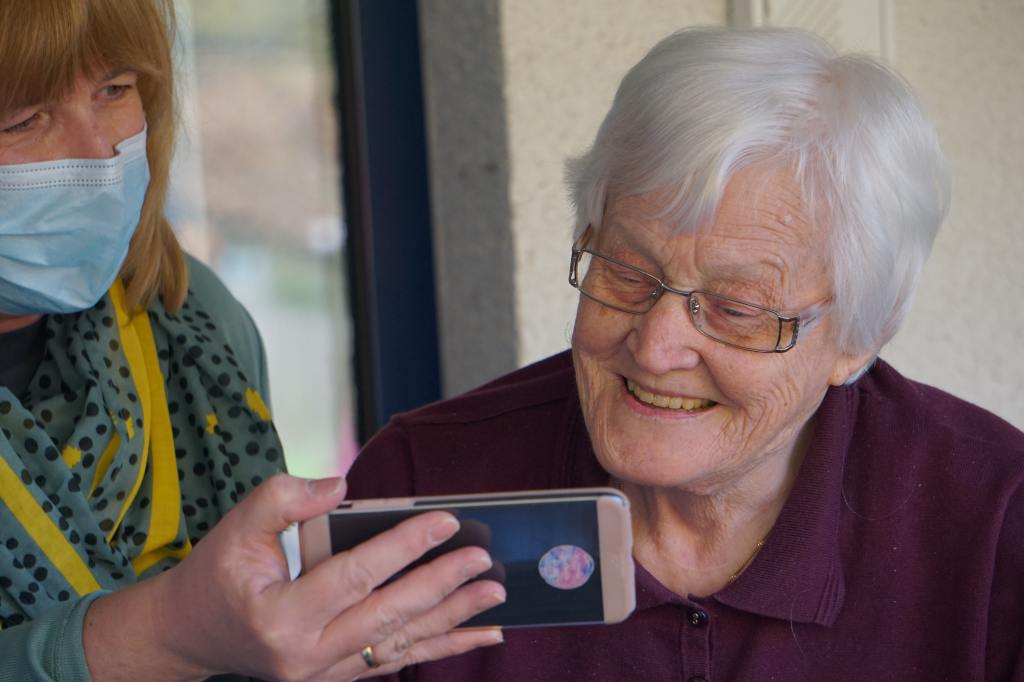 Woman in facemask and green shirt holding up a smartphone to show an elderly woman, who is looking at the phone and smiling.